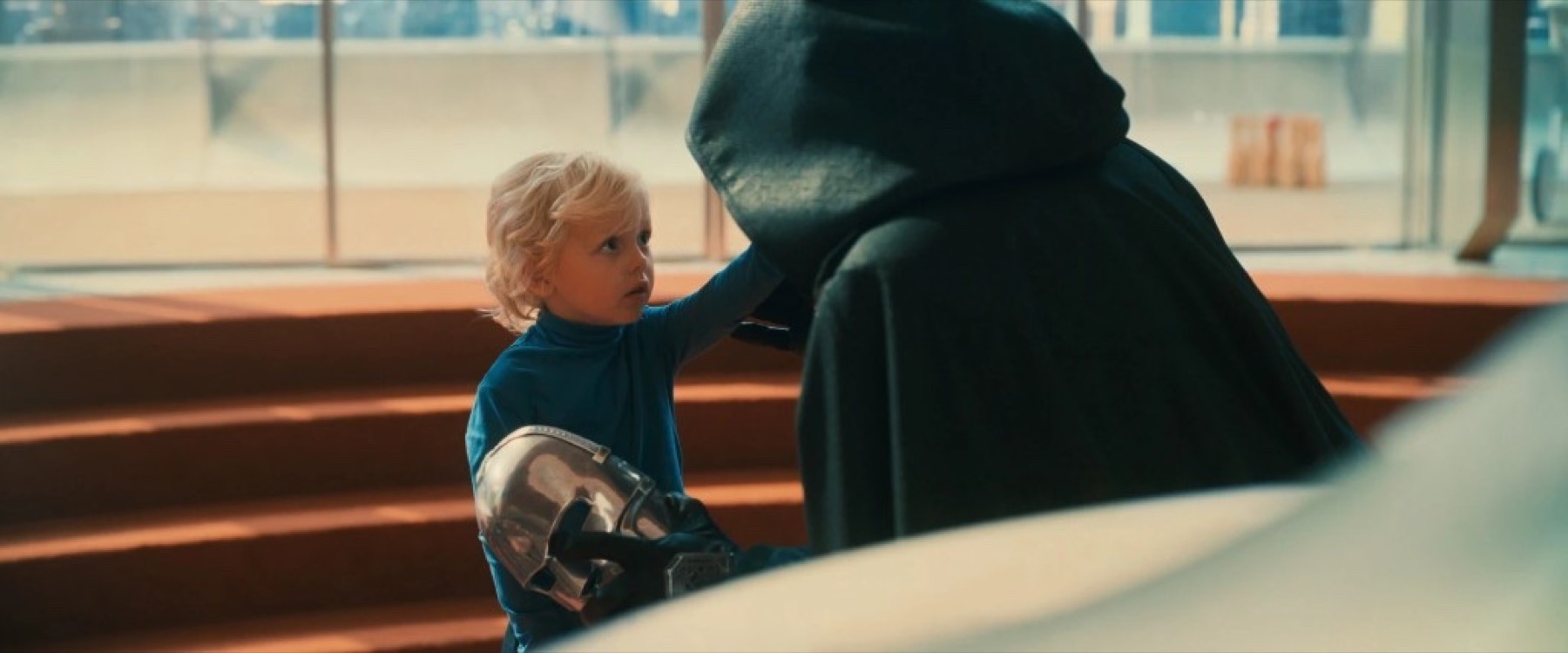 A child holds Doctor Doom’s steel mask beside a figure in a green hooded cloak